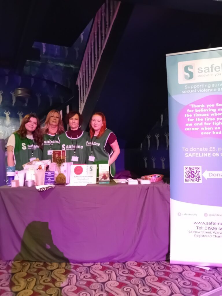 Four women stood behind a stall at the Japan Festival with a range of items on. All of them are wearing green tabbards on with Safeline's logo.