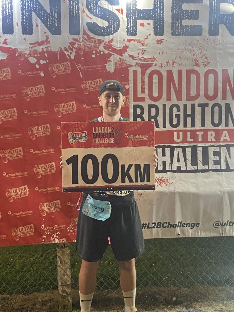Fundraiser holding a banner saying 100km after completing the London 2 Brighton Ultra Challenge