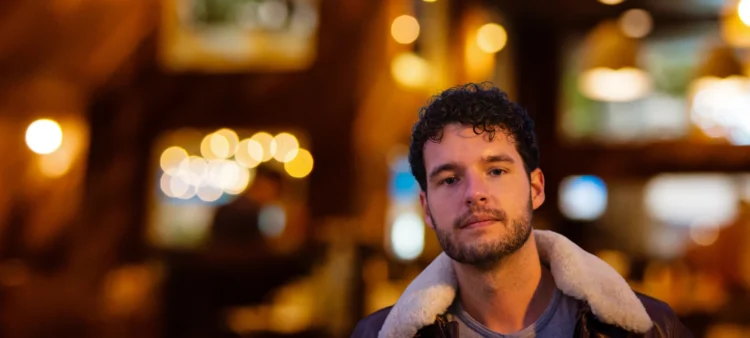 Young man in his twenties with a close cropped beard and moustache. Dark curly hair. Wearing a jacket with a fur collar.