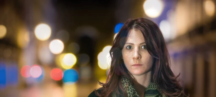Young woman with shoulder length brown hair and hazel eyes is looking at you. Behind her are street lights blurred