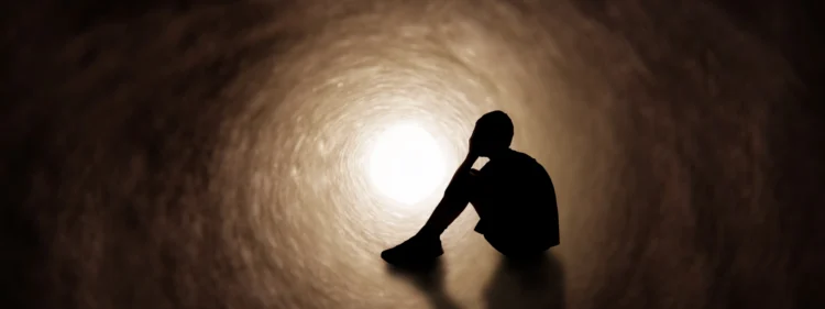 Silhouette of a person with head in hands. sitting in a dark tunnel illuminated by light at the far end.